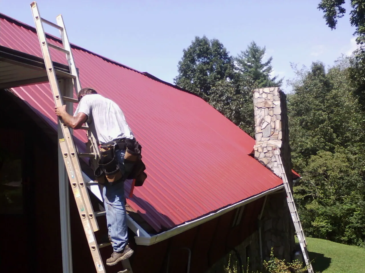 Roofer on a ladder beside a red metal roof and stone chimney — Soffit & Fascia Repair by Roofing Brothers in Leavenworth